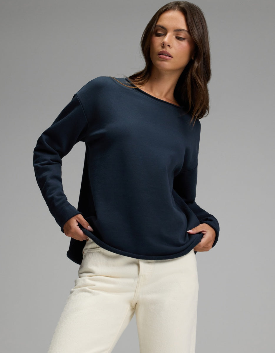 A woman wearing the Cozy Earth Women’s Chelsea Pullover in Eclipse and off-white pants stands against a plain gray background, looking to her left with her hands in her pockets.