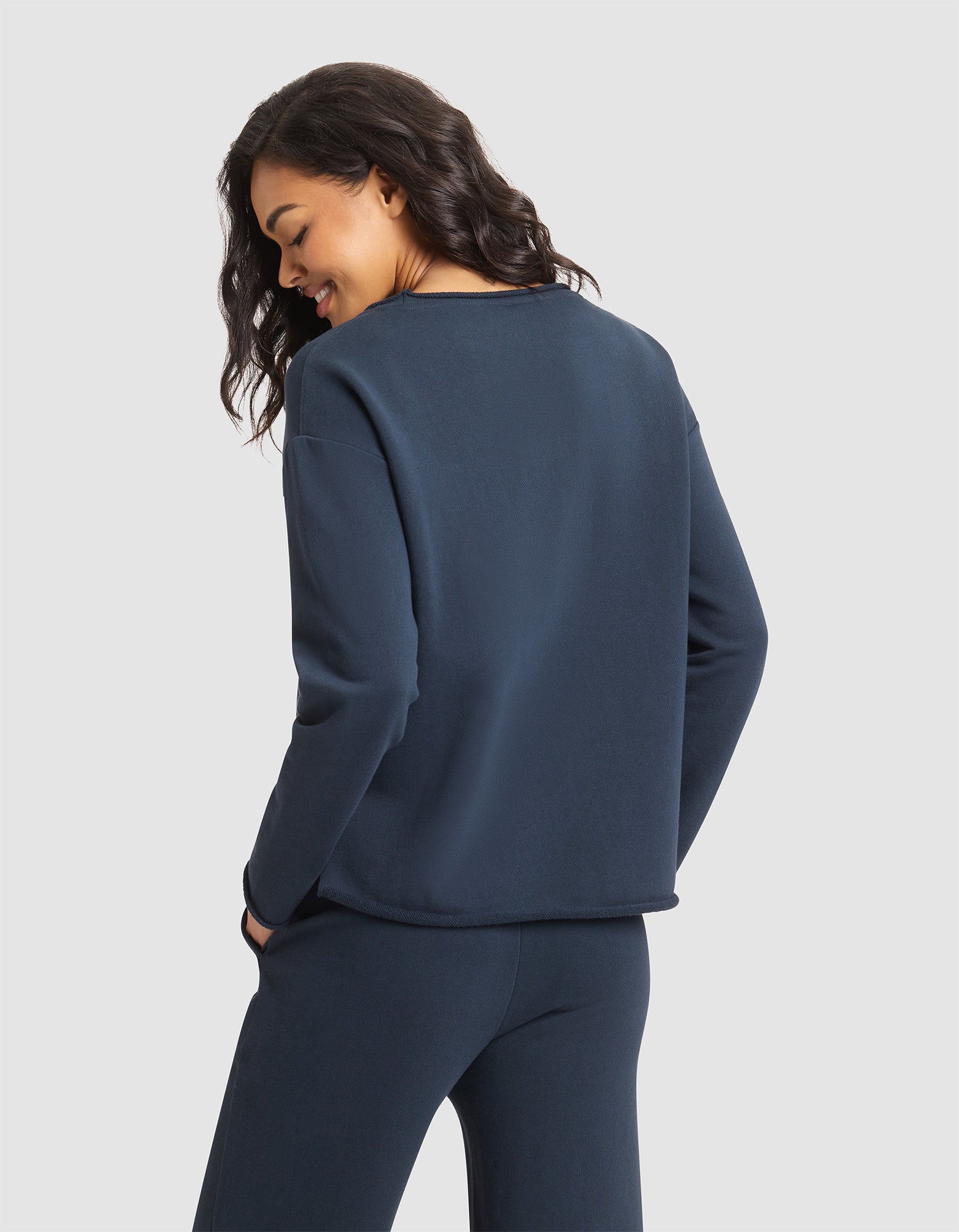 A woman with wavy dark hair, wearing the Cozy Earth Women's Chelsea Pullover in navy blue and matching pants, stands against a plain white background, facing away and smiling over her shoulder. 