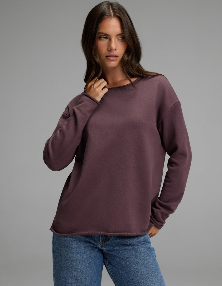 A woman wearing the Cozy Earth Women’s Chelsea Pullover in Fig and blue jeans stands against a plain gray background, looking slightly to the side with one hand near her neck.|Color:Fig