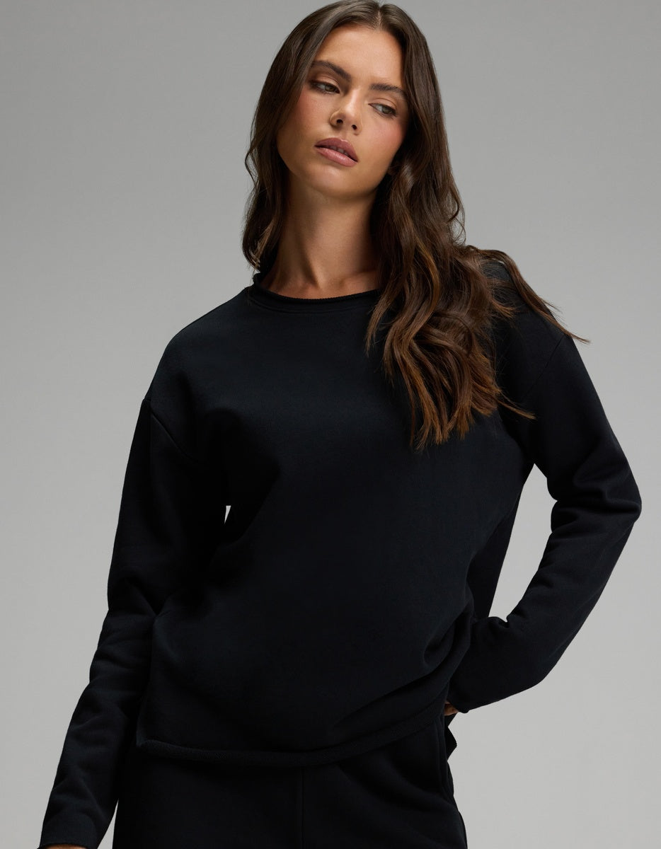 A woman with long brown hair is wearing the Cozy Earth Women’s Chelsea Pullover in Jet Black, paired with black pants. She stands with one hand on her hip, looking to the side against a plain gray background.