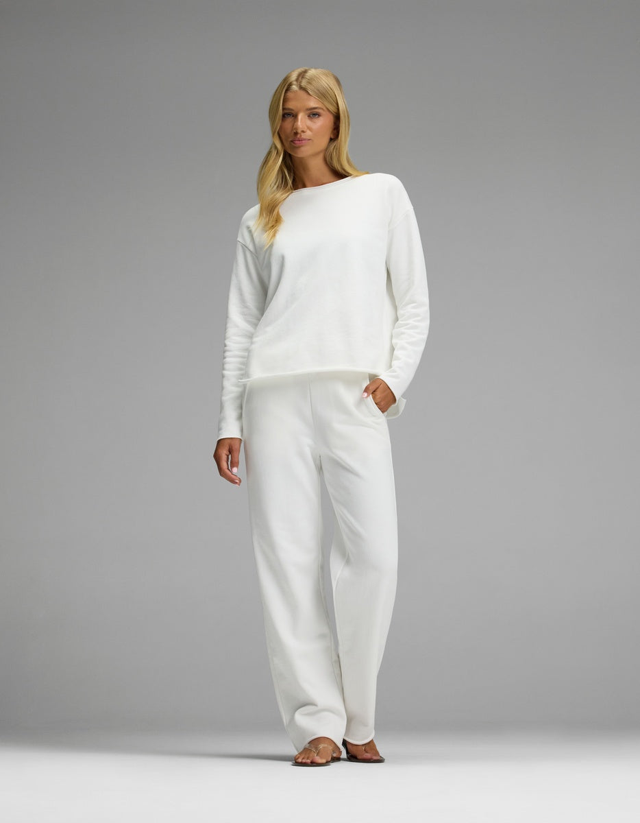 A woman with long blonde hair stands barefoot against a plain gray background, wearing the Women’s Chelsea Set— a loose-fitting white long-sleeve top and matching white pants from Cozy Earth.