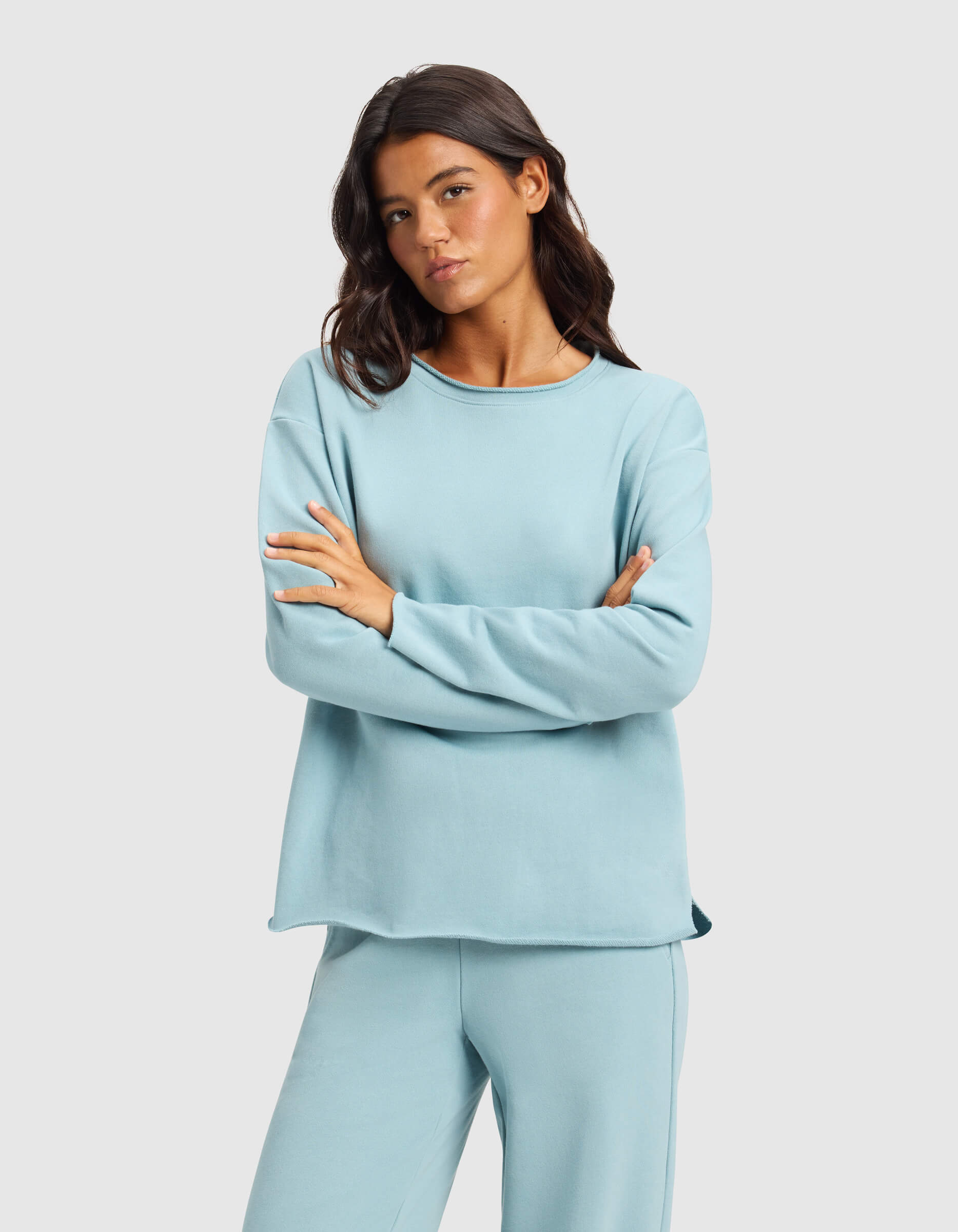 A woman with long dark hair stands with arms crossed, wearing the Cozy Earth Women's Chelsea Pullover and matching pants in light blue against a plain light gray background.|Color:River