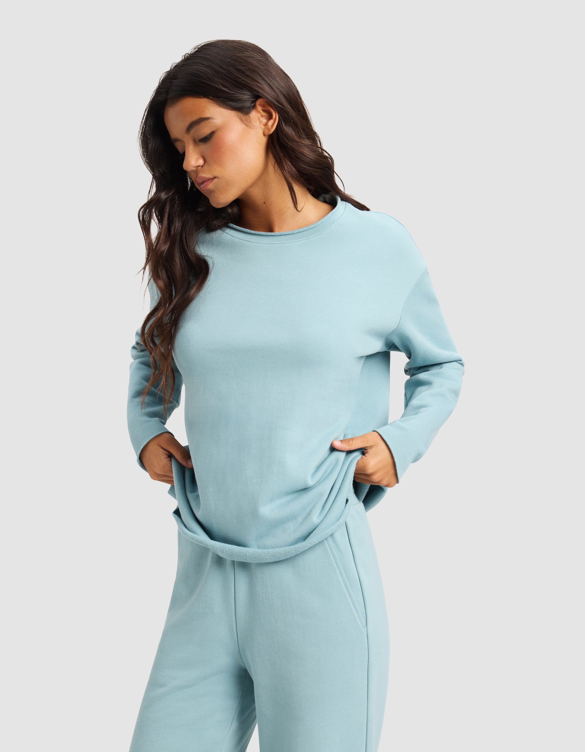 A woman with long brown hair wears the Cozy Earth Women's Chelsea Pullover in light blue and matching sweatpants, standing against a plain light gray background with her hands in her pockets, looking down.|Color:River