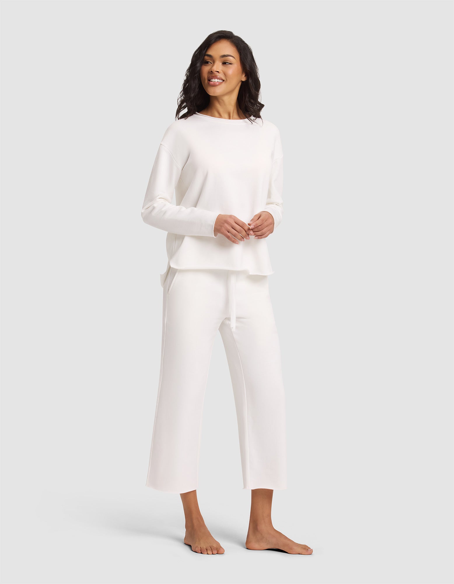 A woman stands barefoot, smiling, wearing the Cozy Earth Women's Chelsea Pullover—a loose-fitting, long-sleeve white top—paired with matching wide-leg pants against a plain light gray background. 