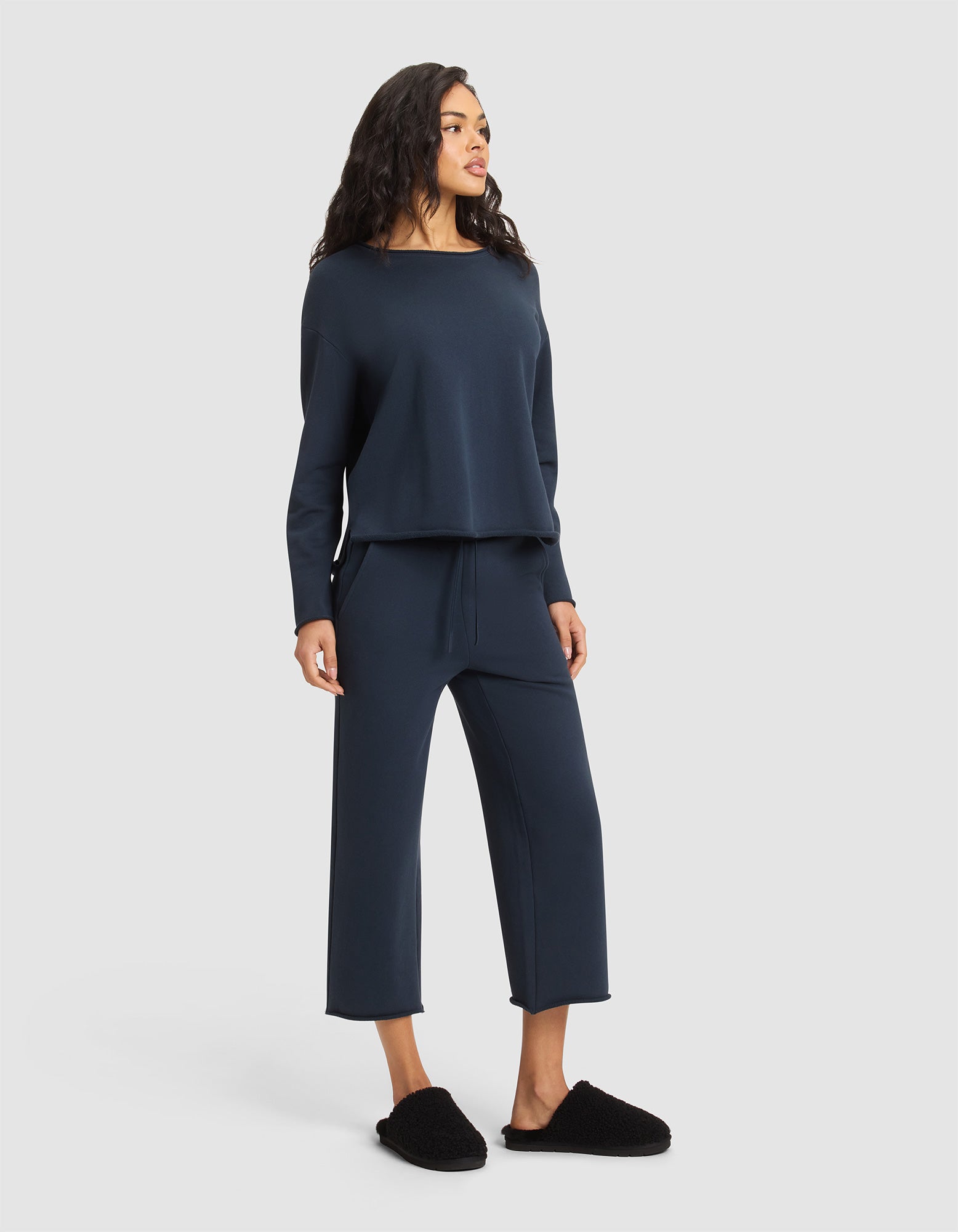 A woman faces slightly left in a loose-fitting navy Women's Chelsea Pullover and matching wide-leg pants from Cozy Earth, paired with black slippers, standing against a plain light background. 