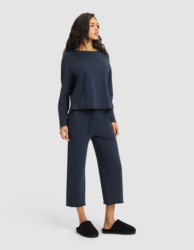A woman faces slightly left in a loose-fitting navy Women's Chelsea Pullover and matching wide-leg pants from Cozy Earth, paired with black slippers, standing against a plain light background.