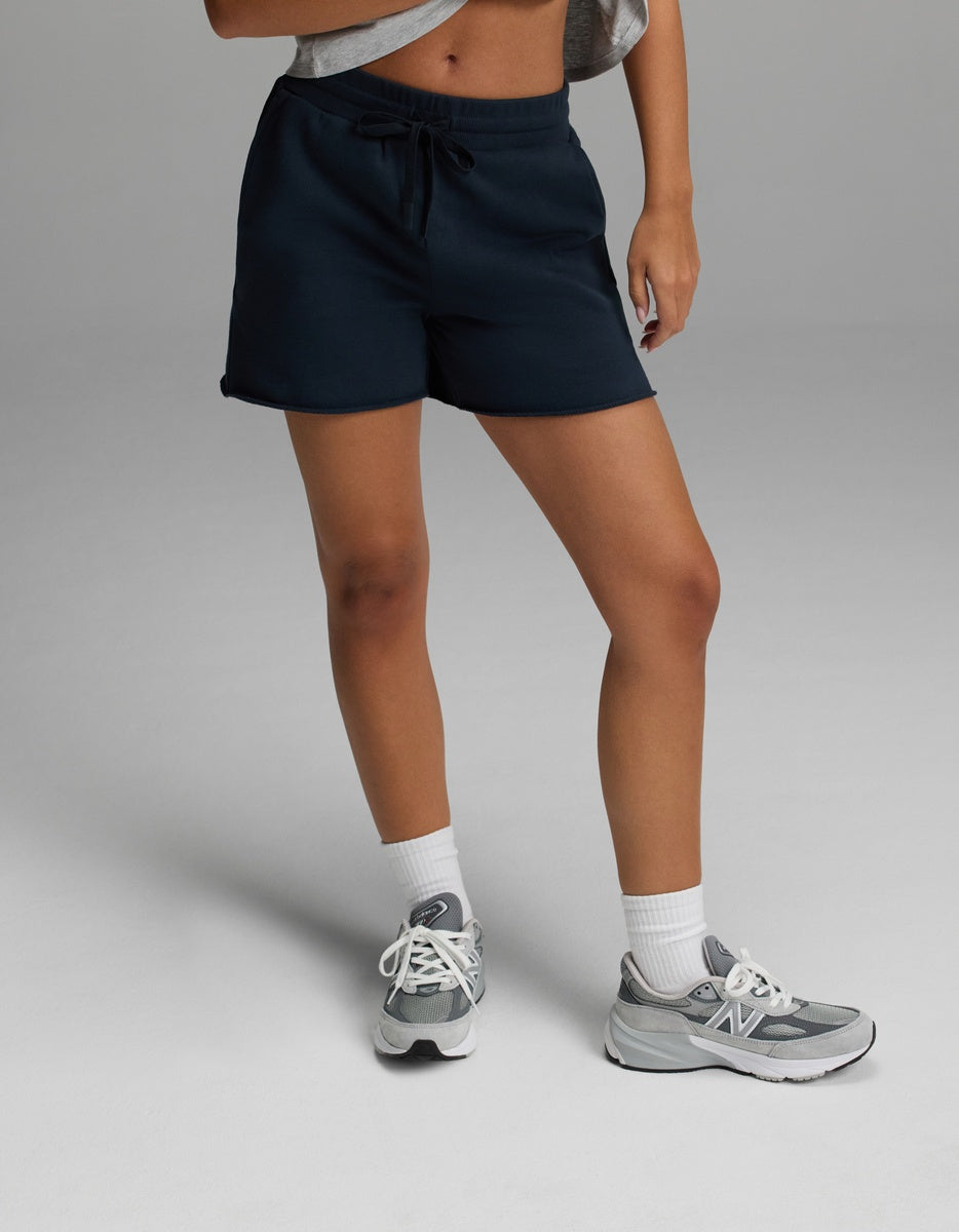 A person wearing the Cozy Earth Women’s Chelsea Short in Eclipse, a gray cropped top, white socks, and gray New Balance sneakers stands on a light gray surface. Their face is not visible in the image.