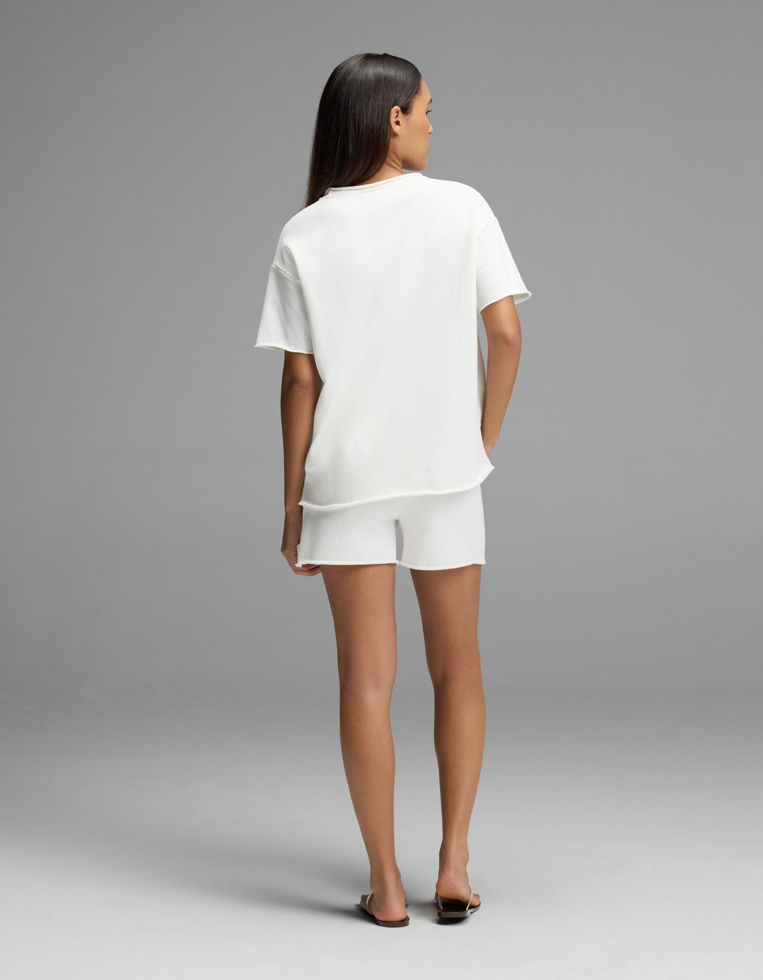 A woman stands facing away in the Cozy Earth Women’s Chelsea Short Sleeve Set, featuring a loose white t-shirt and matching white shorts, paired with black sandals against a plain gray background.