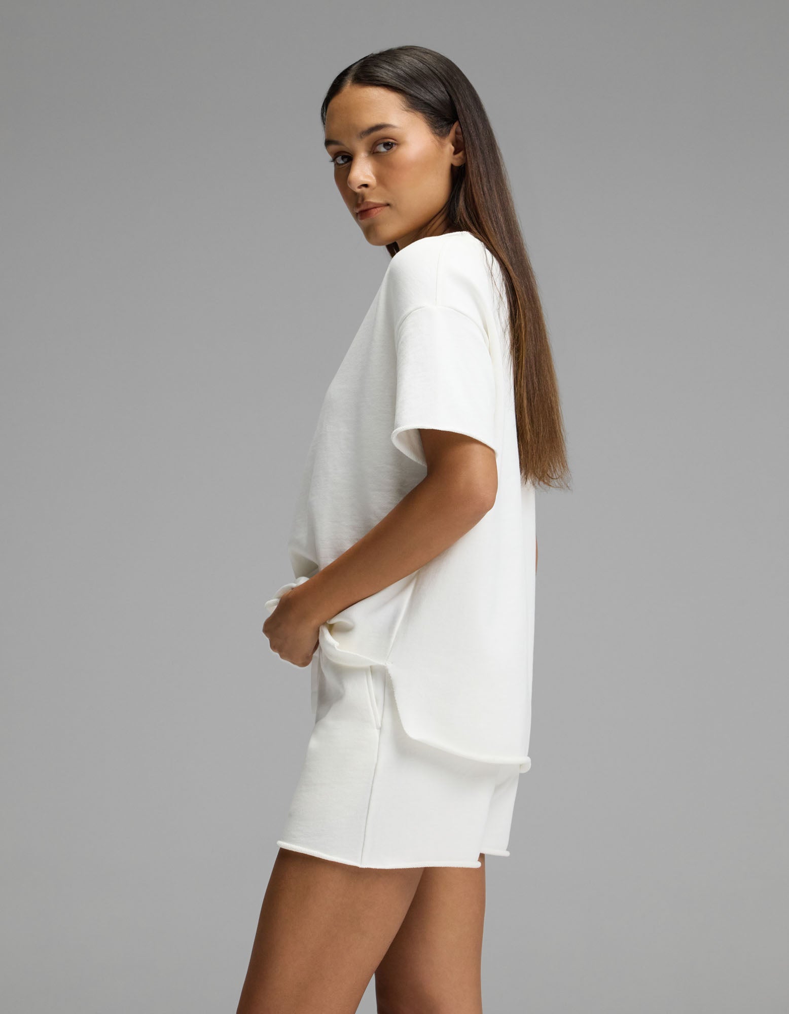 A woman with long straight hair stands sideways against a plain gray background, hands in the pockets of her Cozy Earth Women’s Chelsea Short Sleeve Set—a loose white T-shirt paired with matching white shorts.