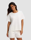 A woman with long dark hair models the Women's Chelsea Short Sleeve by Cozy Earth, paired with white shorts. She stands against a light gray background in a relaxed, casual pose with a slight smile.|Color:Bone