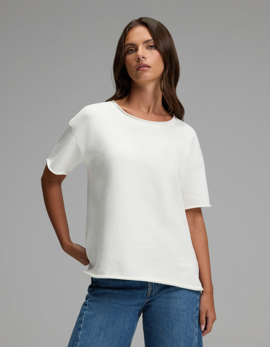 A woman with long brown hair stands against a neutral background, wearing the Cozy Earth Women’s Chelsea Short Sleeve in Bone and blue jeans. She has one hand in her pocket and a confident expression.