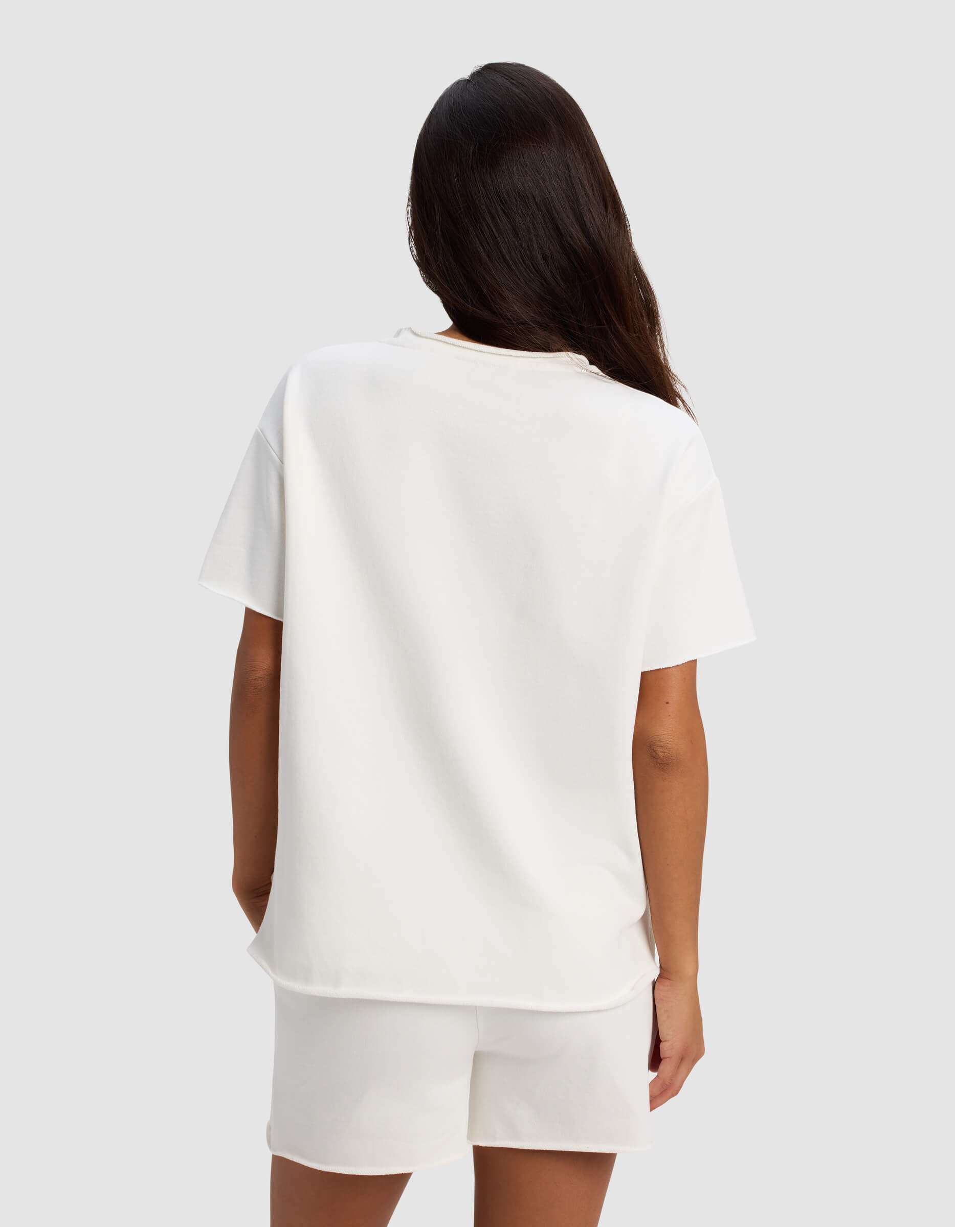 A woman with long dark hair, seen from behind, wears the Cozy Earth Women's Chelsea Short Sleeve and white shorts against a plain light gray background.