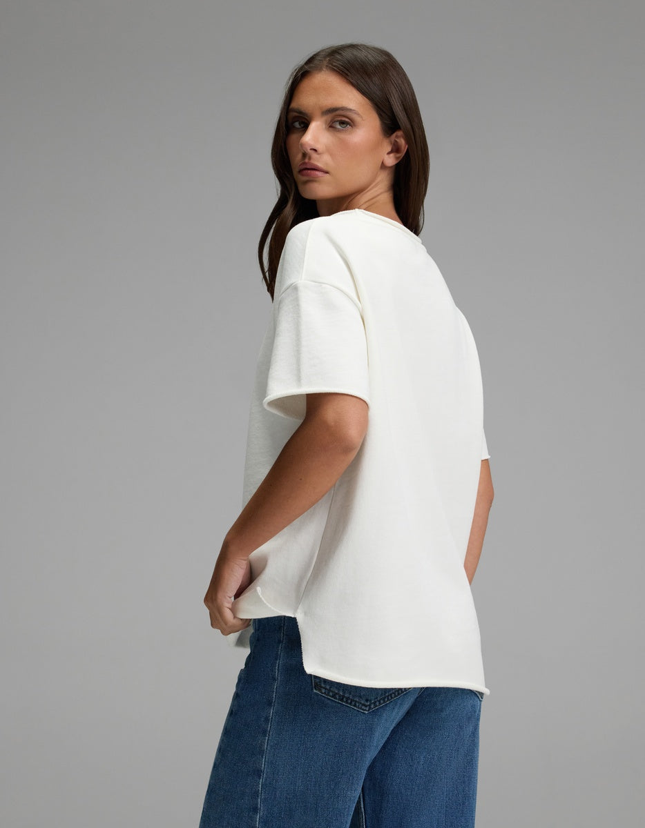 A woman with long brown hair, dressed in the Cozy Earth Women’s Chelsea Short Sleeve and blue jeans, stands against a plain gray background, glancing over her shoulder at the camera.