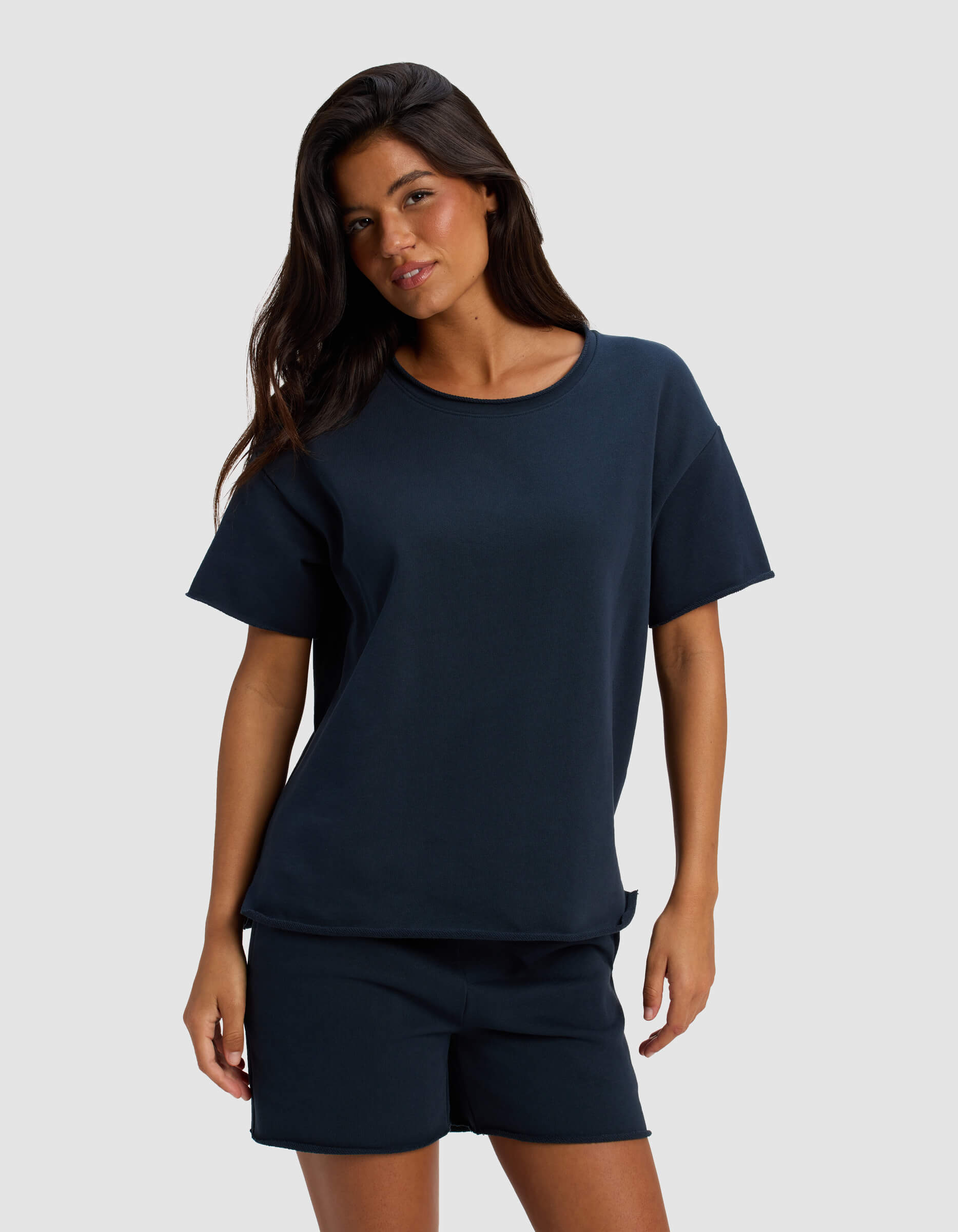 A woman with long dark hair wears Cozy Earth's Women's Chelsea Short Sleeve navy blue t-shirt and matching shorts, standing against a plain light gray background.