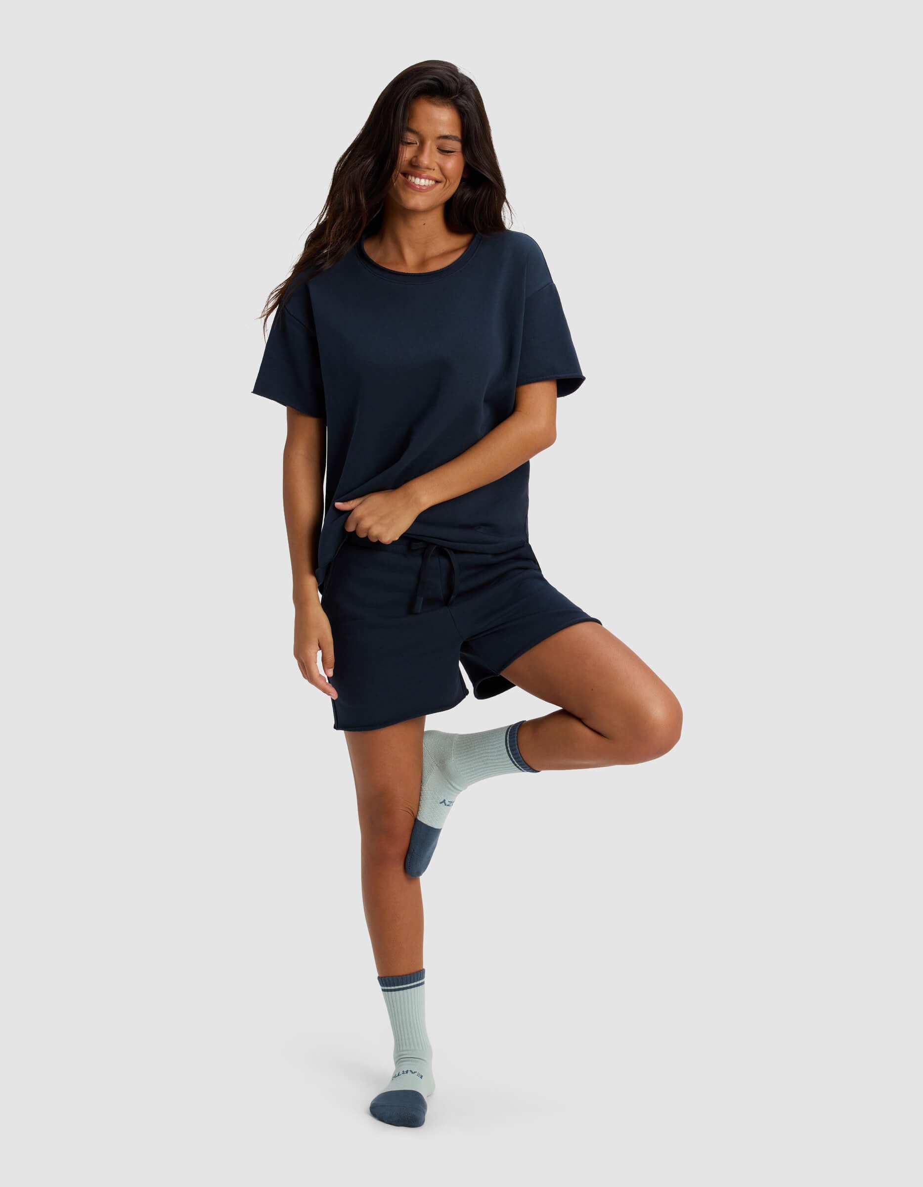 A woman stands on one leg, smiling with her hands by her sides, wearing Cozy Earth's Women's Chelsea Short Sleeve in navy blue and matching shorts, plus light blue socks with dark blue accents, against a plain light gray background.
