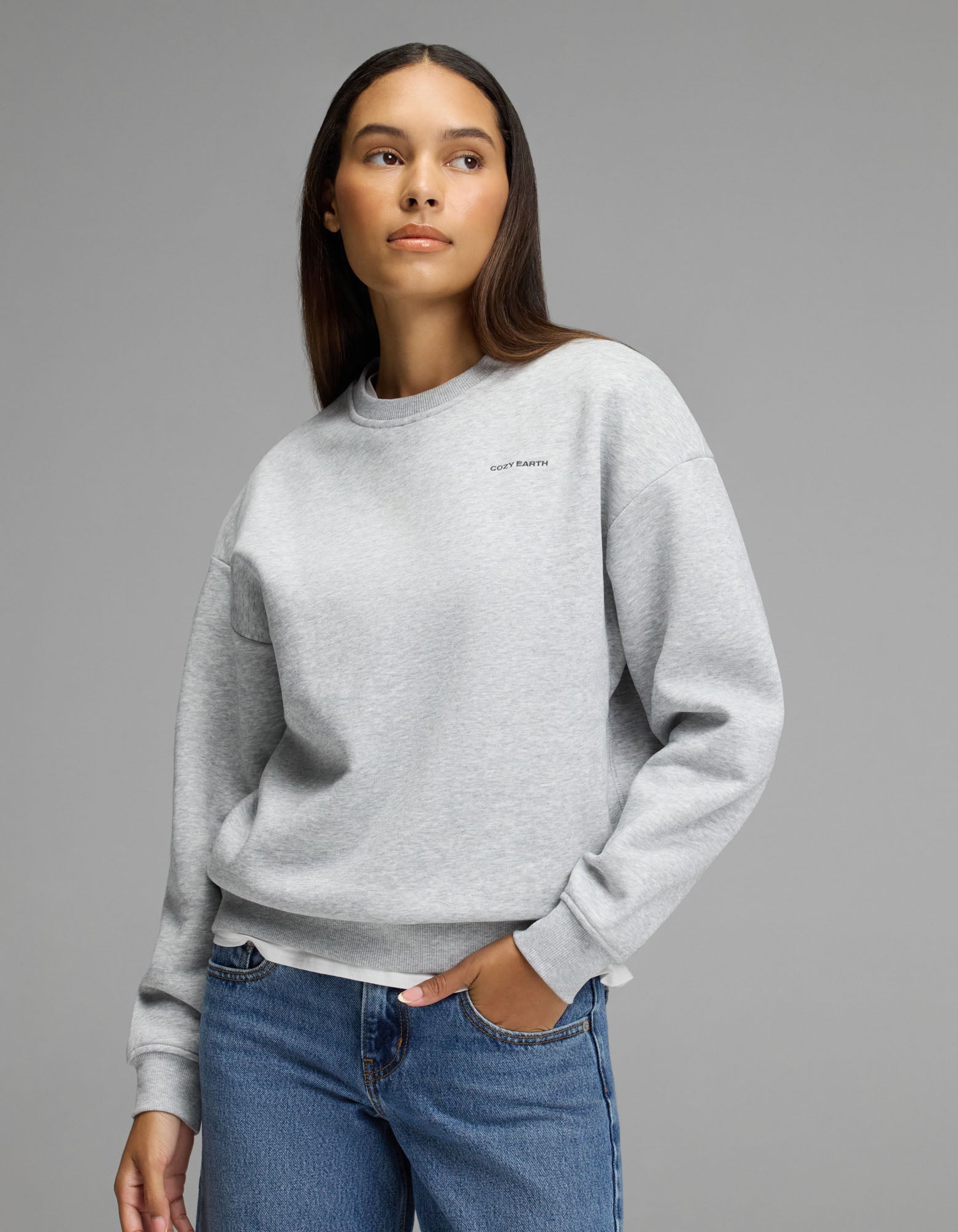 A person models the Cozy Earth Women’s Cityscape Crewneck in Heather Grey, paired with blue jeans, standing against a plain gray background and glancing to the side with one hand in their pocket.