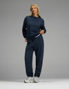 A woman with blonde hair stands confidently against a plain gray background, wearing the Cozy Earth Women’s CityScape Crewneck & Sweatpant Set in navy blue, paired with white sneakers.|Color:Eclipse