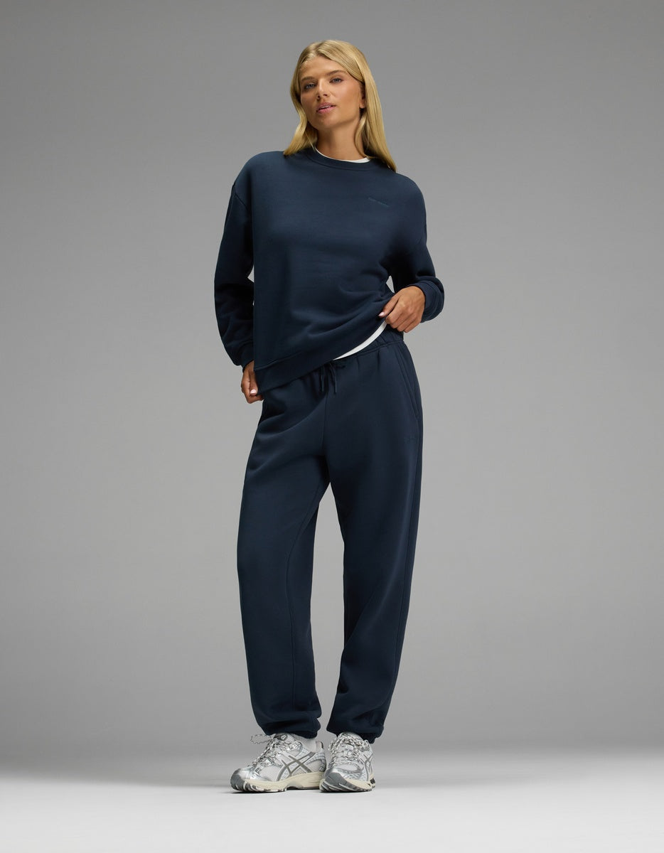 A woman with blonde hair stands confidently against a plain gray background, wearing the Cozy Earth Women’s CityScape Crewneck & Sweatpant Set in navy blue, paired with white sneakers.