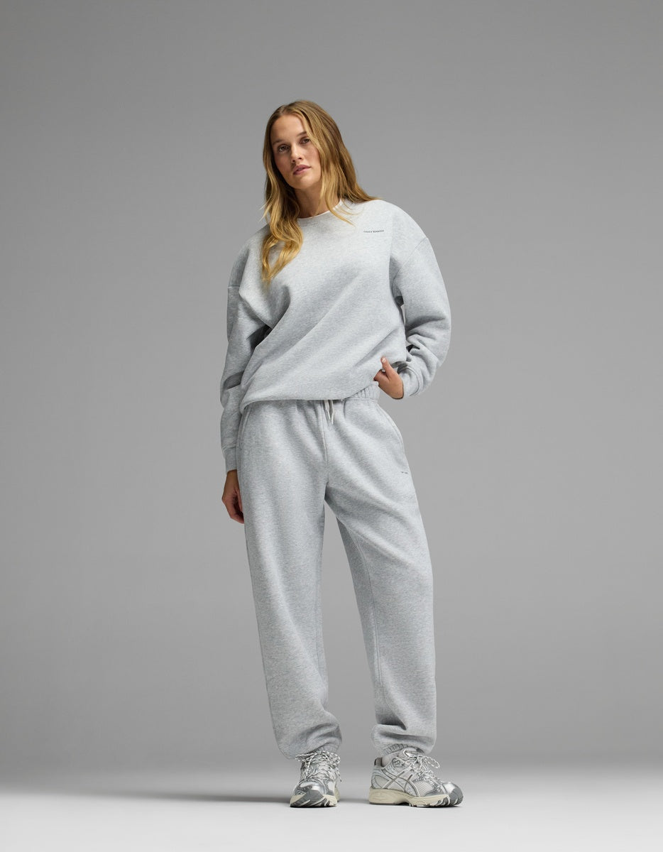 A woman stands against a plain gray background wearing the Cozy Earth Women’s CityScape Crewneck & Sweatpant Set and silver athletic sneakers, with one hand in her pocket.|Color:Heather Grey