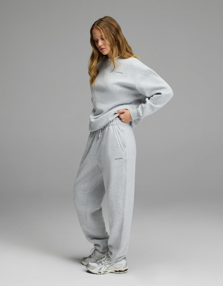 A woman stands in profile wearing the Cozy Earth Women’s CityScape Crewneck & Sweatpant Set in light gray, paired with gray sneakers. She has long, light brown hair and looks down with one hand in her pocket against a plain gray background.|Color:Heather Grey
