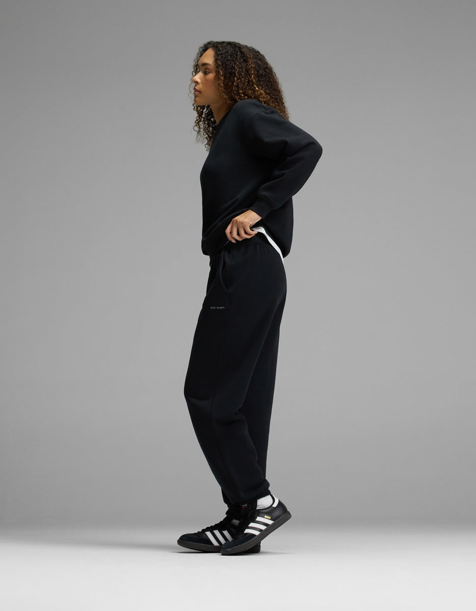 A person with curly hair stands sideways in a relaxed pose, wearing the Women’s CityScape Crewneck & Sweatpant Set by Cozy Earth in black, paired with black Adidas sneakers featuring white stripes, against a plain gray background.|Color:Jet Black