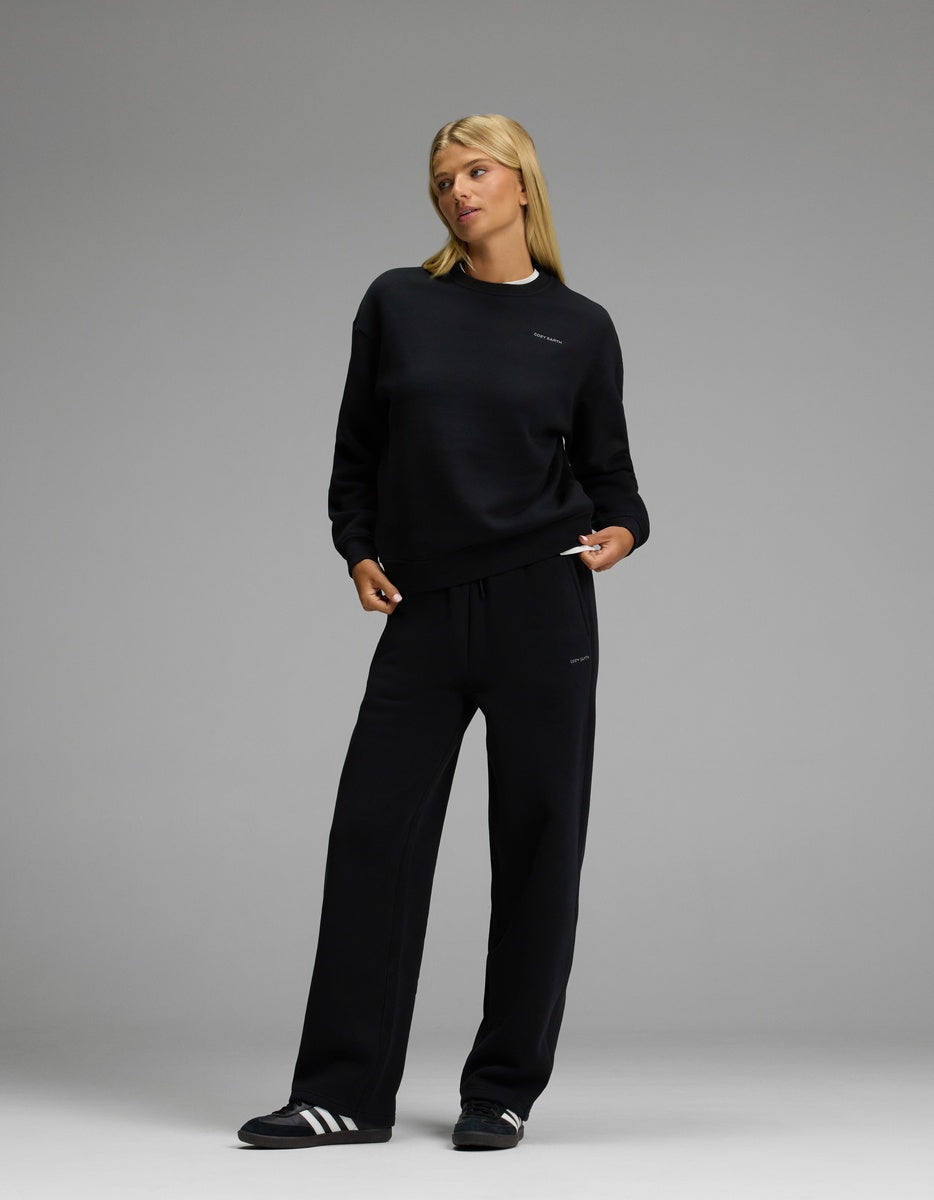 A person wearing the Cozy Earth Women’s CityScape Crewneck & Wide Leg Pant Set in black stands against a plain gray background, looking slightly to the side. They complete the look with black and white athletic shoes.|Color:Jet Black