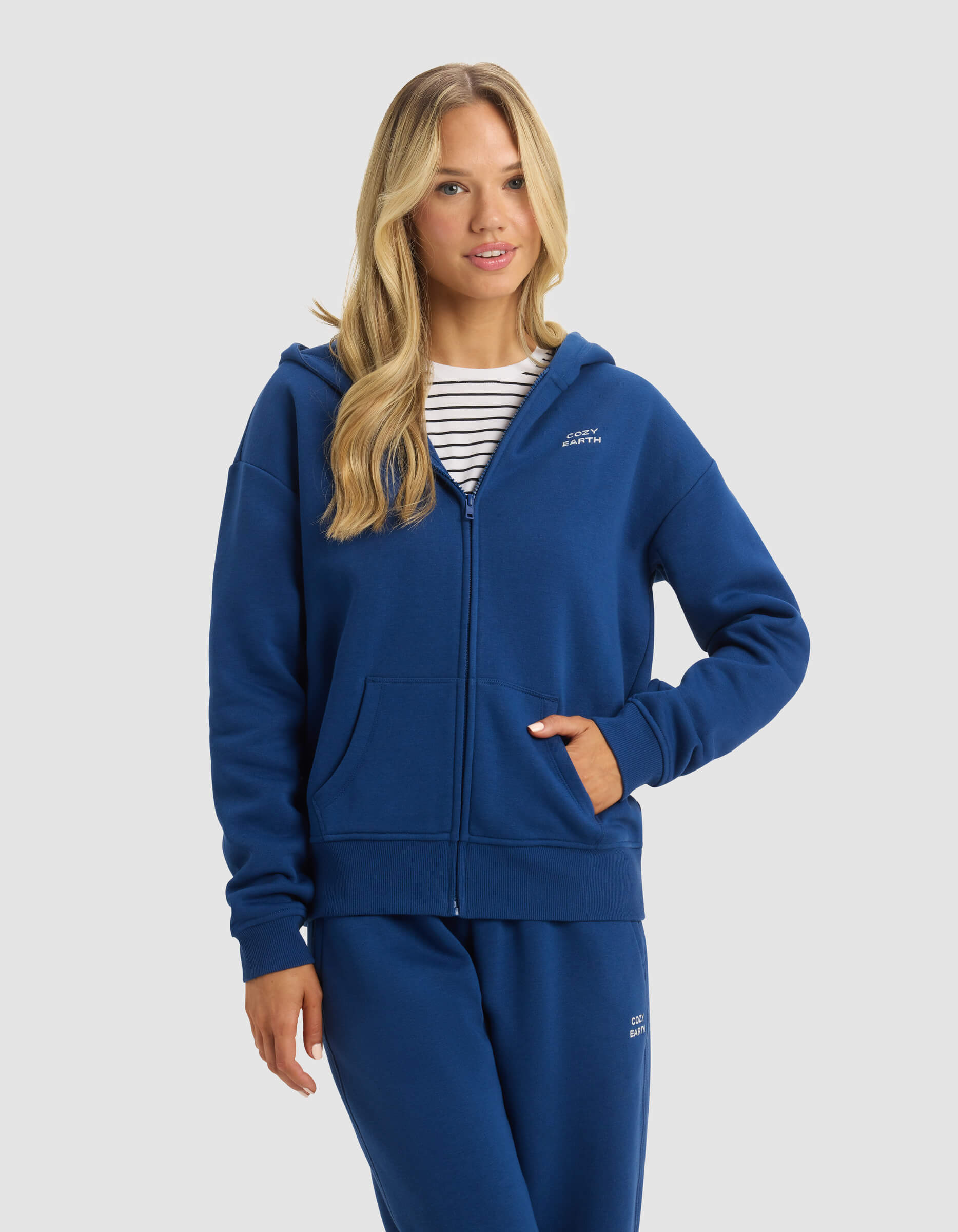 A woman with long blonde hair models the Cozy Earth Women's CityScape Full Zip hoodie and matching blue sweatpants over a white and navy striped shirt, posing with one hand in her pocket against a plain light background.|Color:Pacific