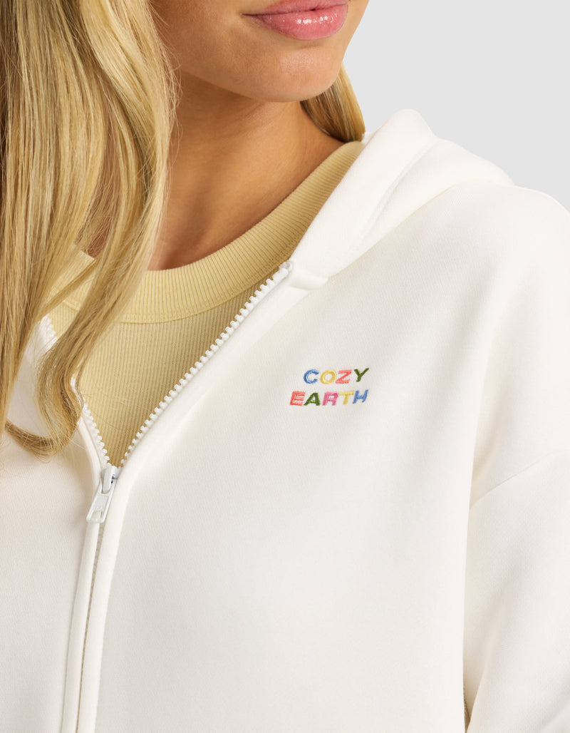 A woman wears the Cozy Earth Women's CityScape Full Zip—a white hoodie with colorful "COZY EARTH" embroidery on the chest—over a beige top. Only her lower face, neck, and part of her blonde hair are visible.
