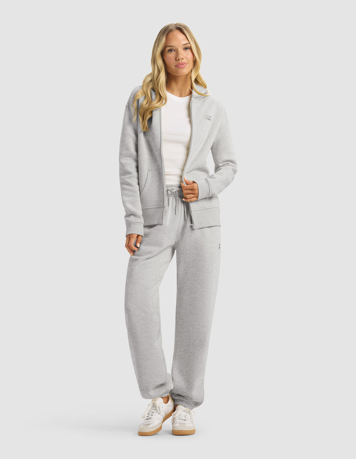 A woman with long blonde hair wears the Cozy Earth Women's CityScape Full Zip Hoodie & Sweatpant Set in light gray, paired with a white t-shirt and sneakers, standing relaxed against a plain light background.|Color:Heather Grey