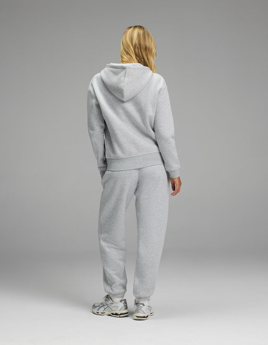A woman with blond hair wears the Women’s CityScape Full-Zip Hoodie & Sweatpant Set by Cozy Earth, standing with her back to the camera against a plain gray background and wearing white sneakers.