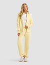 A woman with long blonde hair wears the Cozy Earth Women's CityScape Full Zip & Sweatpant Set in light yellow, paired with a white t-shirt and sneakers. She stands facing forward against a plain light gray background.|Color:Meringue
