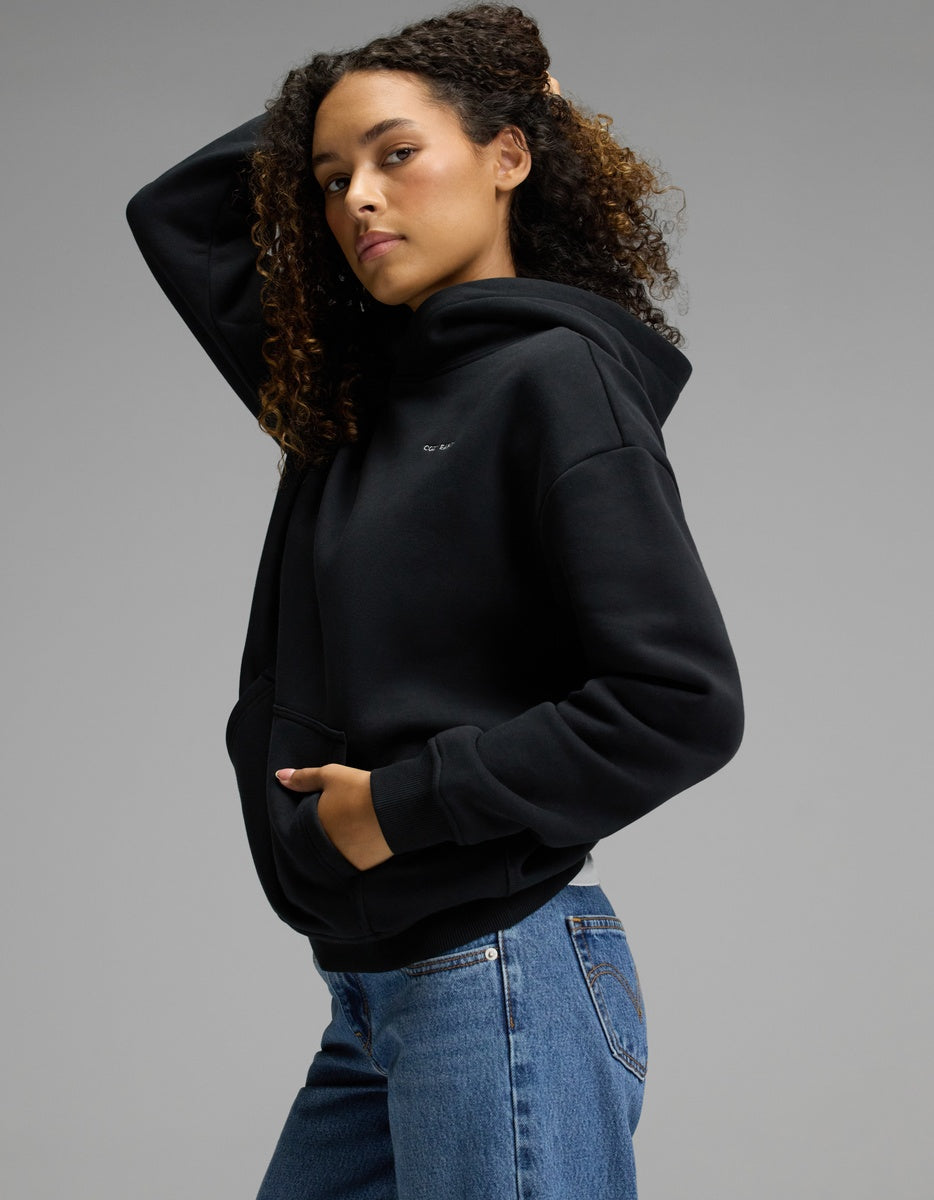 A person with curly hair poses in the Cozy Earth Women’s CityScape Hoodie in Jet Black and blue jeans against a plain gray background, one hand in the hoodie pocket and the other touching their hair.