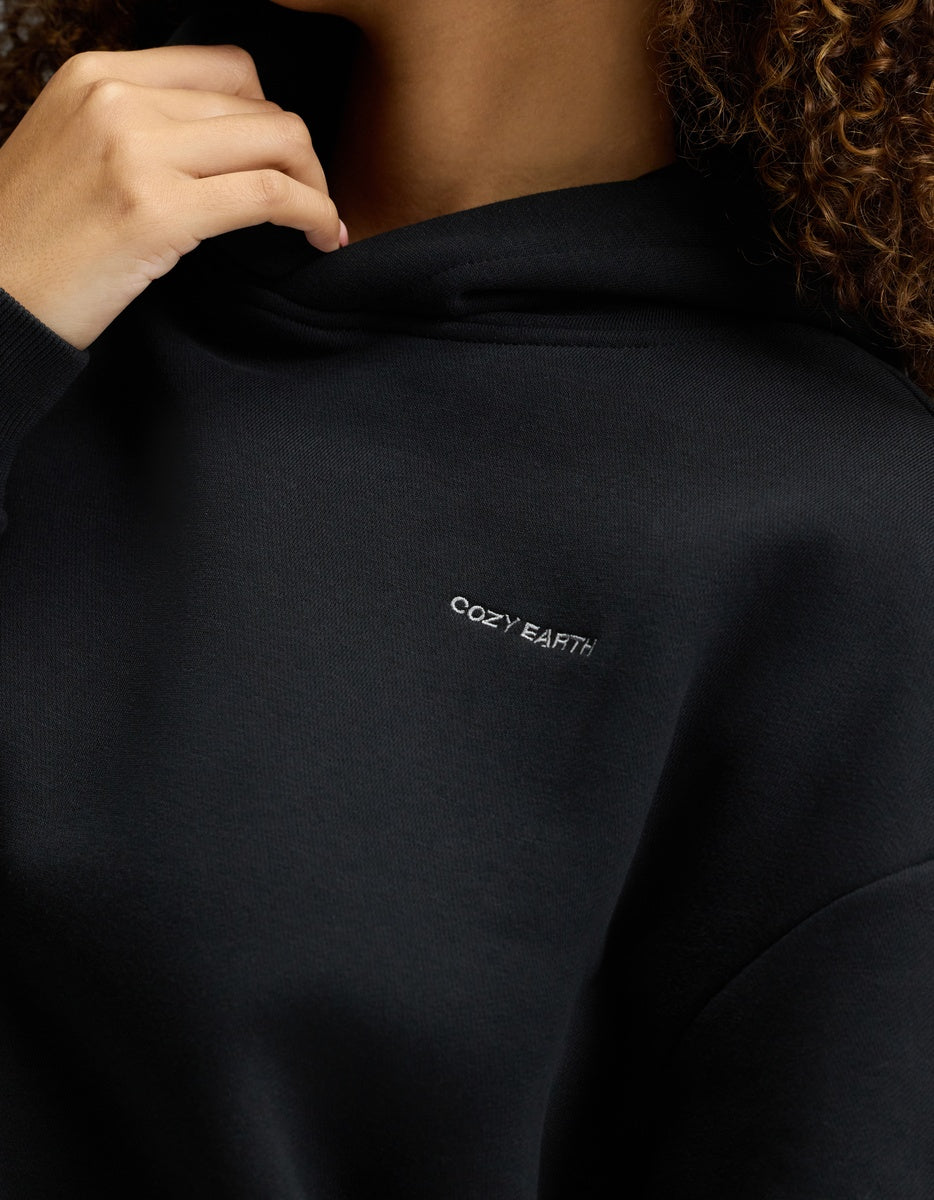 A person wearing the Cozy Earth Women’s CityScape Hoodie in black, featuring “COZY EARTH” embroidered in white on the chest, holds the hood near their face with only part of their face and hand visible.|Color:Jet Black