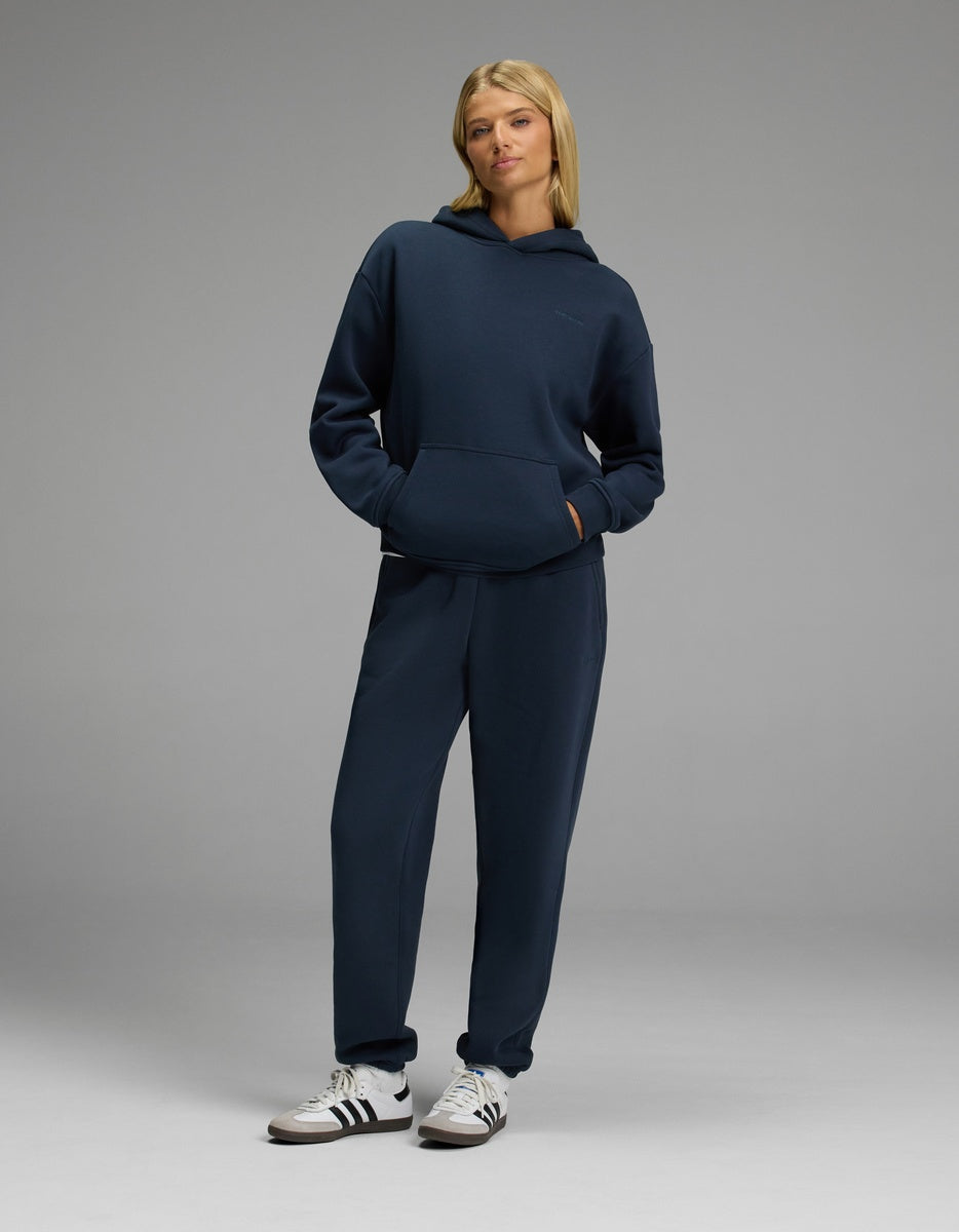 A woman stands in a relaxed pose wearing the Cozy Earth Women’s CityScape Hoodie & Sweatpant Set in navy blue, paired with white sneakers against a plain gray background.|Color:Eclipse