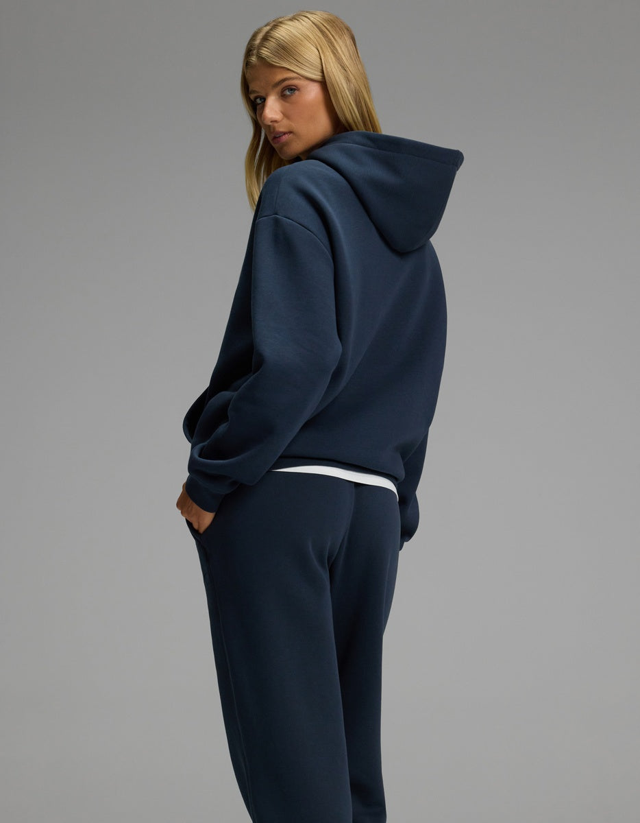 A woman with long blonde hair models the Cozy Earth Women’s CityScape Hoodie & Sweatpant Set in navy blue, standing sideways and looking over her shoulder against a plain gray background.|Color:Eclipse