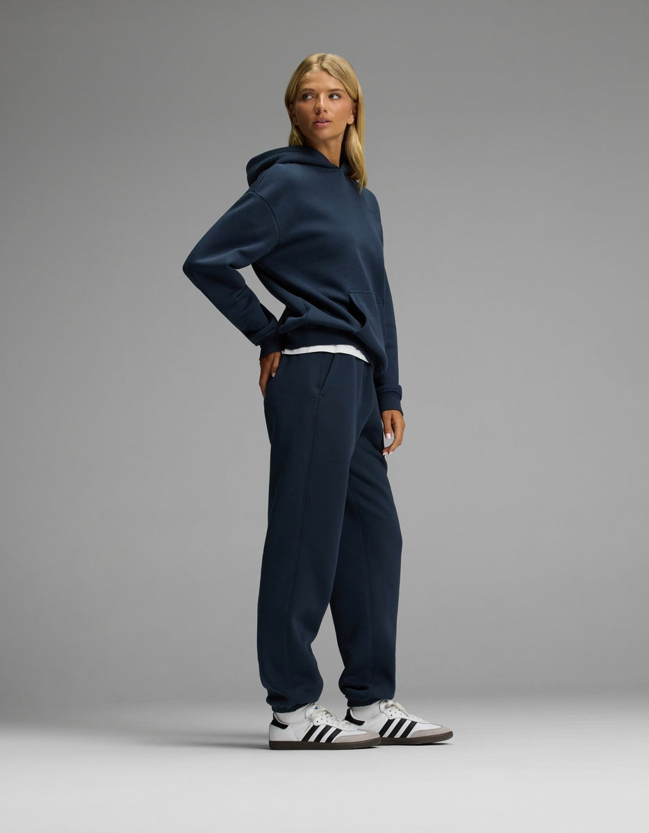 A person stands in a relaxed pose wearing the Cozy Earth Women’s CityScape Hoodie & Sweatpant Set in navy, paired with white Adidas sneakers featuring black stripes, against a plain gray background.|Color:Eclipse
