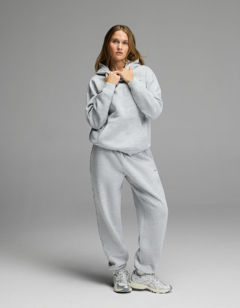 A woman stands against a grey background wearing the Cozy Earth Women’s CityScape Hoodie & Sweatpant Set, paired with silver sneakers. She holds the strings of her hoodie and looks at the camera.