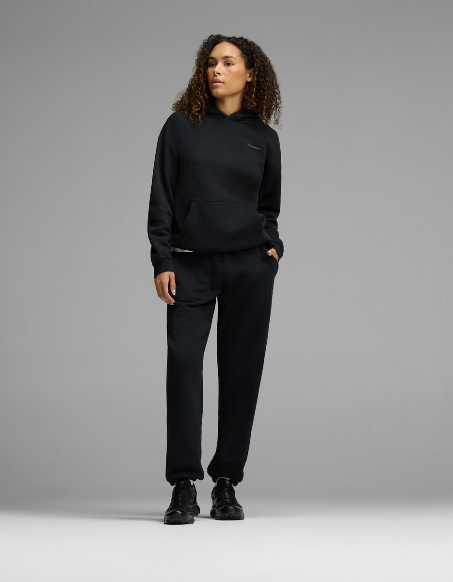 A person with curly hair stands against a gray background, wearing the Cozy Earth Women’s CityScape Hoodie & Sweatpant Set in black, paired with black sneakers and one hand in their pocket.|Color:Jet Black
