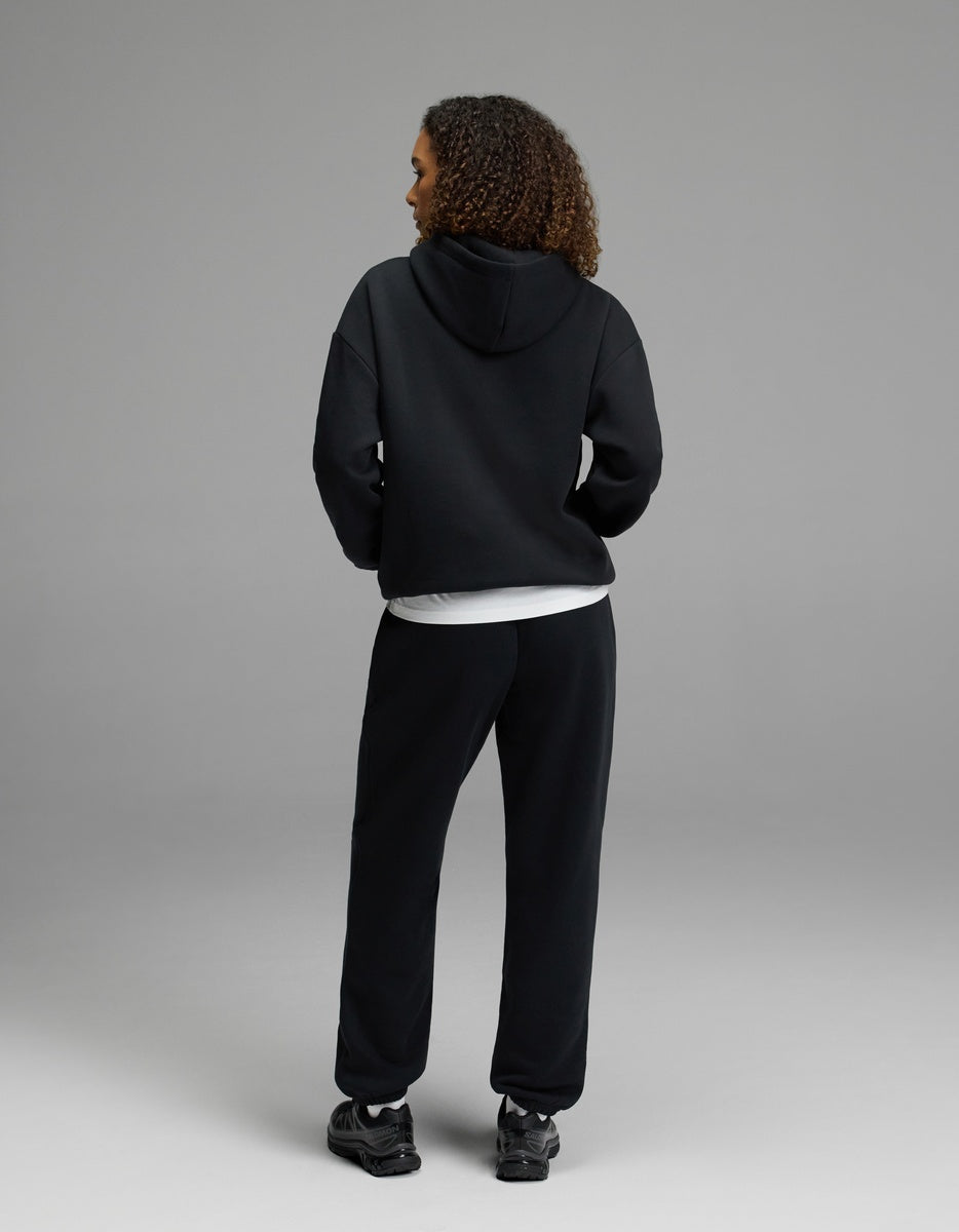 A woman stands with her back to the camera, wearing the black Women’s CityScape Hoodie & Sweatpant Set by Cozy Earth over a white shirt and paired with black sneakers, set against a plain gray background.|Color:Jet Black