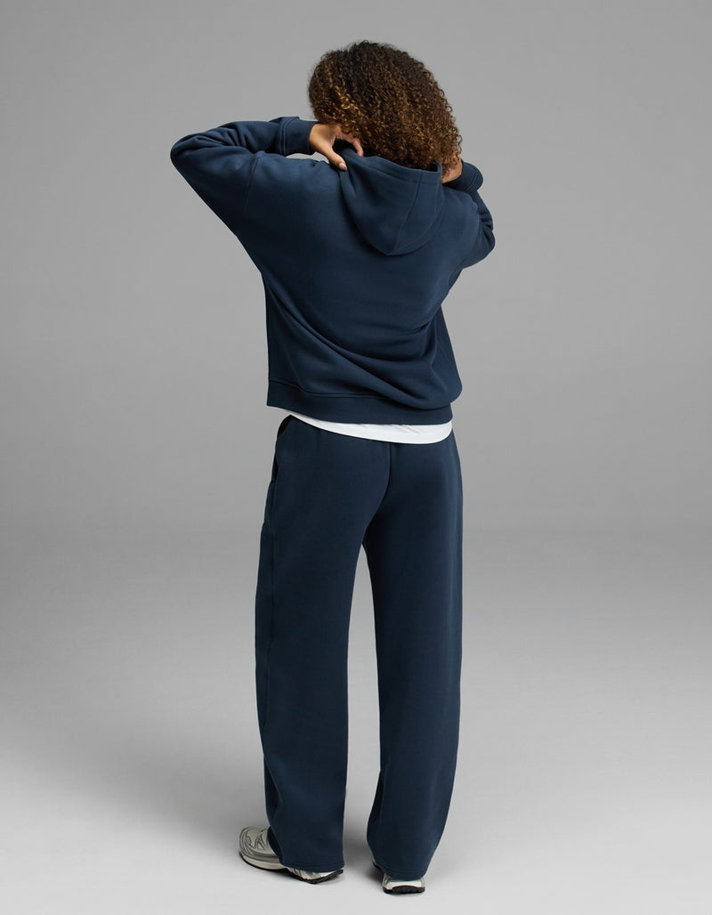 A person with curly hair faces away, adjusting their hood while wearing the Cozy Earth Women’s CityScape Hoodie & Wide Leg Pant Set in navy, paired with white sneakers.