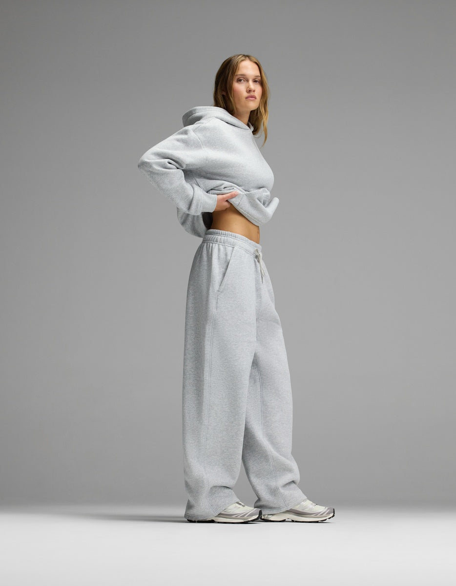 A woman models the Women’s CityScape Hoodie & Wide Leg Pant Set by Cozy Earth in light gray, lifting the hoodie to show her midriff, paired with white sneakers against a plain gray background.