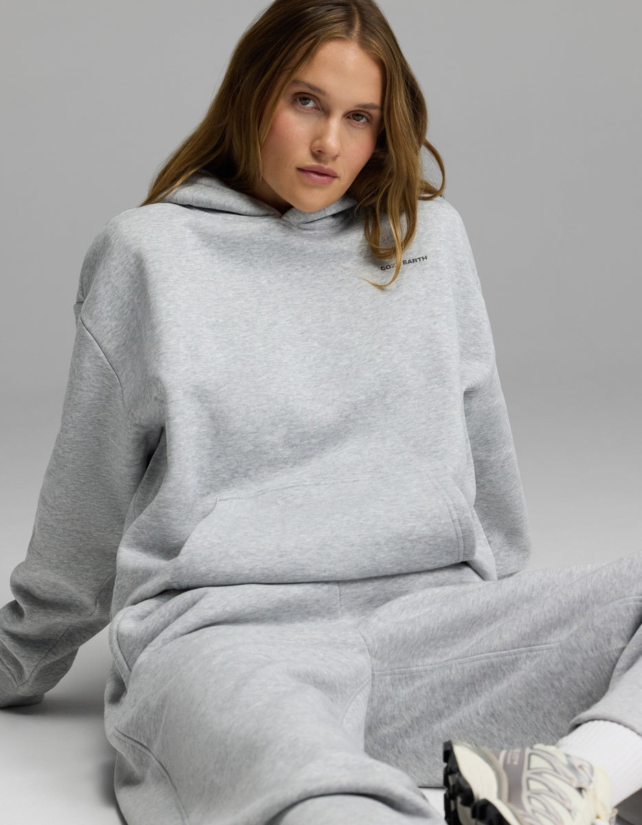 A woman models the Women’s CityScape Hoodie & Wide Leg Pant Set by Cozy Earth, sitting on the floor against a plain gray background and looking at the camera with a relaxed expression.