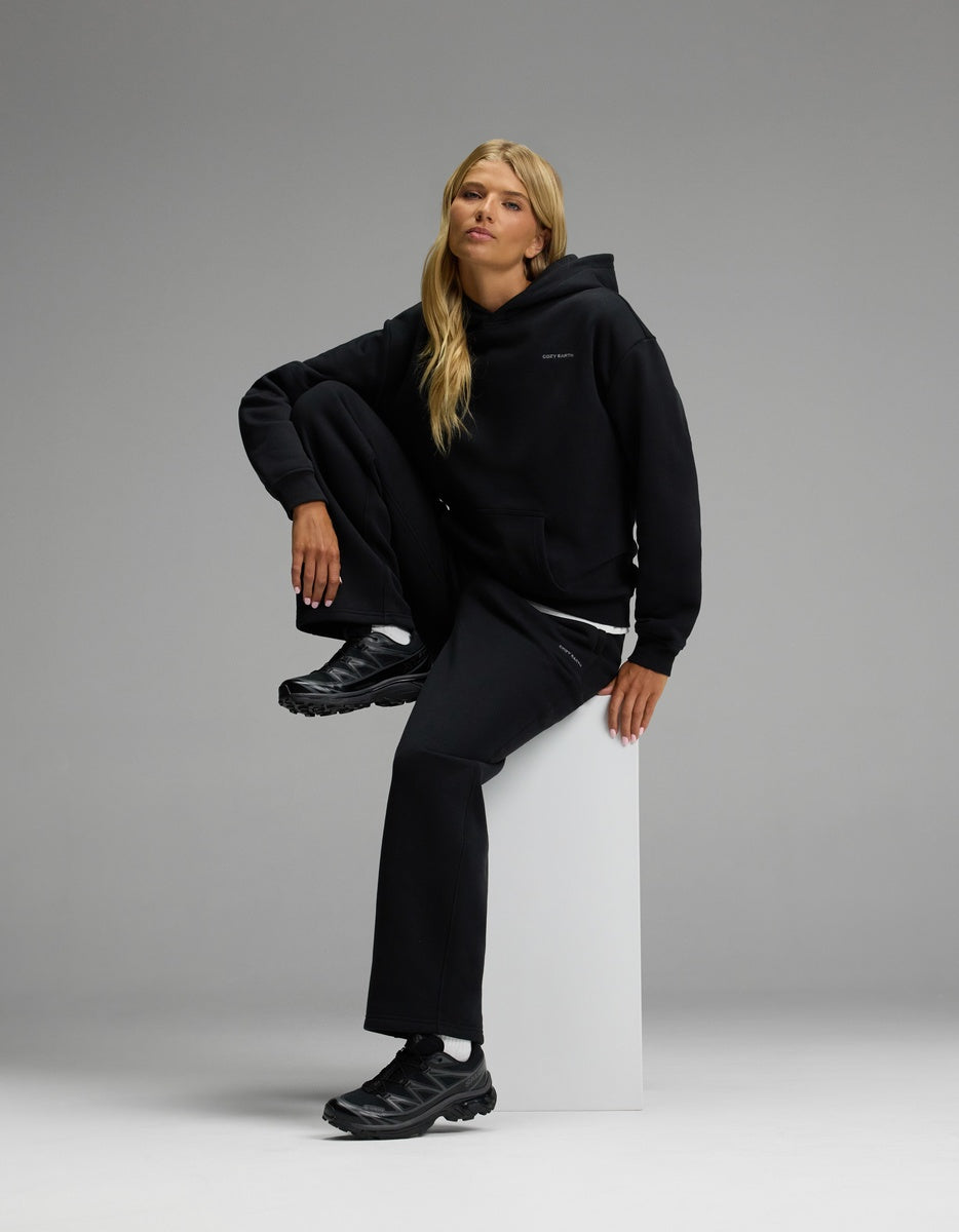 A woman with long blonde hair models the Cozy Earth Women’s CityScape Hoodie & Wide Leg Pant Set in black, seated on a white block against a plain gray background, looking slightly upward.