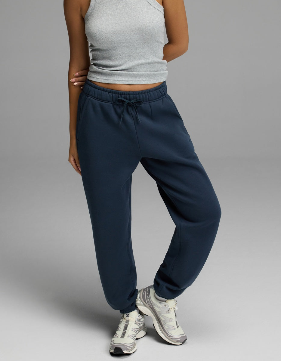 A person is shown from the shoulders down, standing against a plain gray background and wearing navy blue Cozy Earth Women’s CityScape Sweatpants, a grey sleeveless crop top, and white athletic shoes. Their head is not visible in the image.|Color:Eclipse