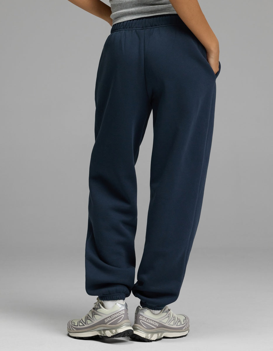 A person stands with their back to the camera, wearing a gray tank top, Cozy Earth Women’s CityScape Sweatpant in navy blue, and gray athletic shoes. One hand is in their pocket against a plain light gray background.|Color:Eclipse