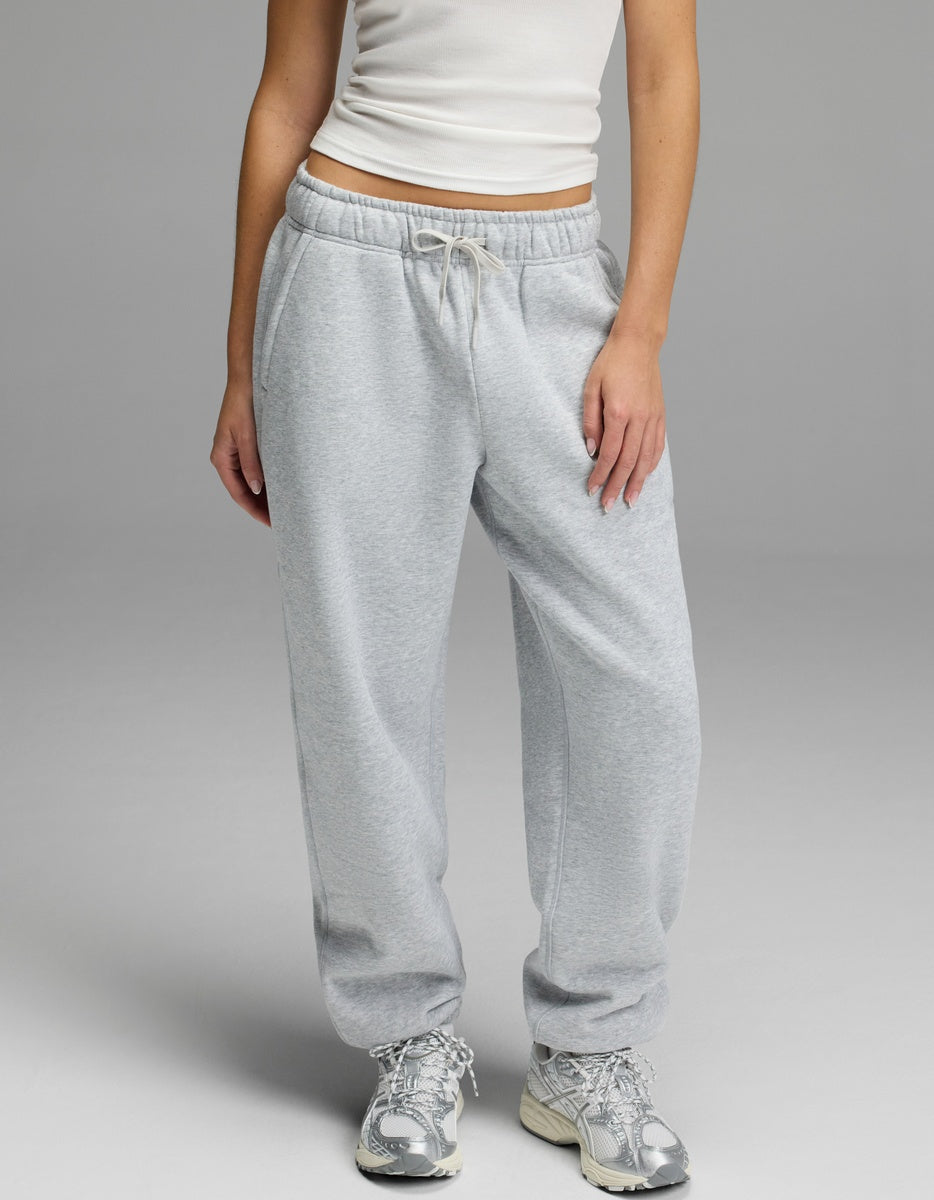 A person models the Cozy Earth Women’s CityScape Sweatpant in light gray, featuring a drawstring waist, paired with a white tank top and silver athletic shoes against a plain gray background. Only the lower half of the body is visible.