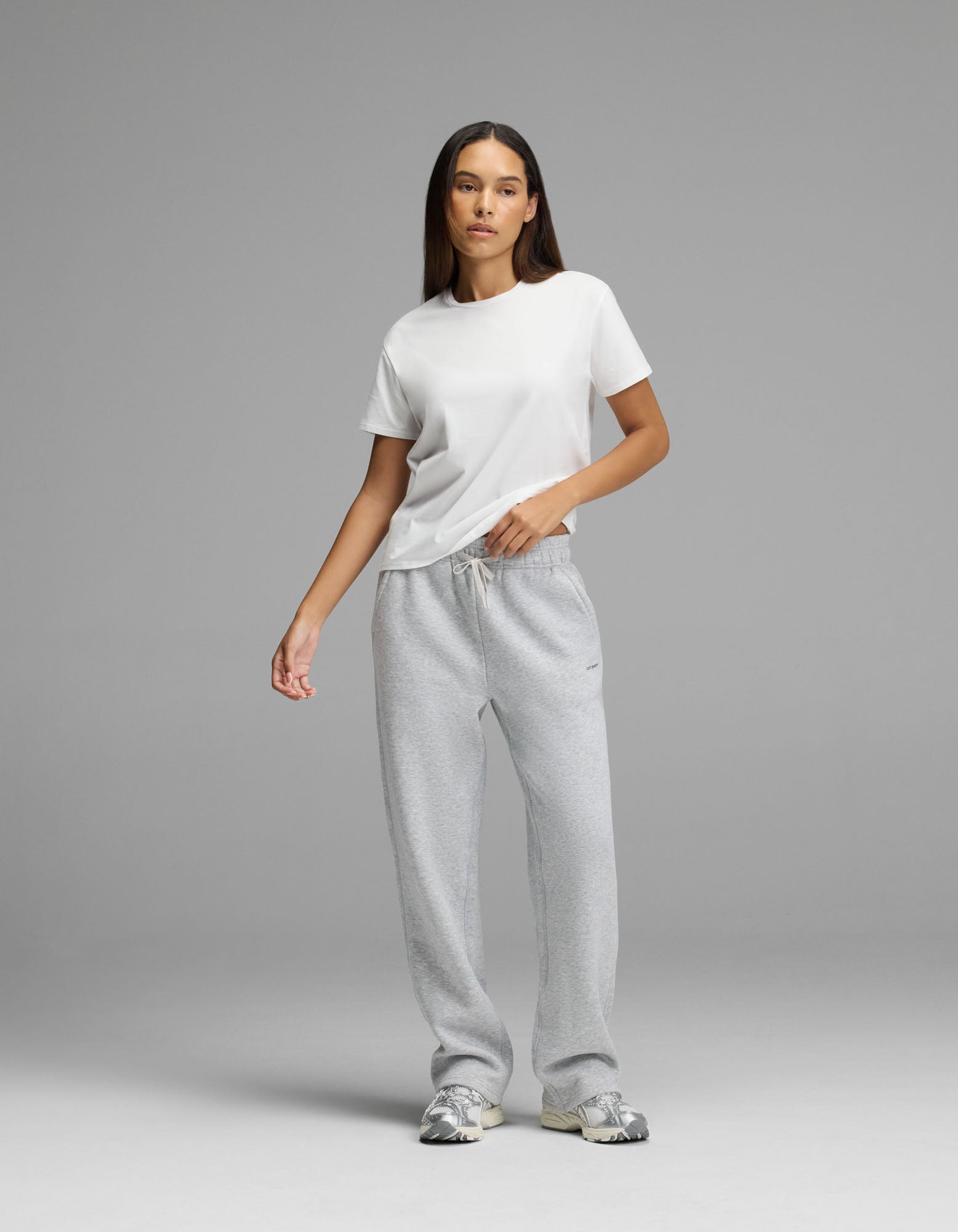 A person stands against a plain gray background wearing a white t-shirt, Cozy Earth Women’s Cityscape Wide Leg Pant in light gray, and gray sneakers. They have long, straight hair and a neutral expression with one hand in their pocket.|Color:Heather Grey