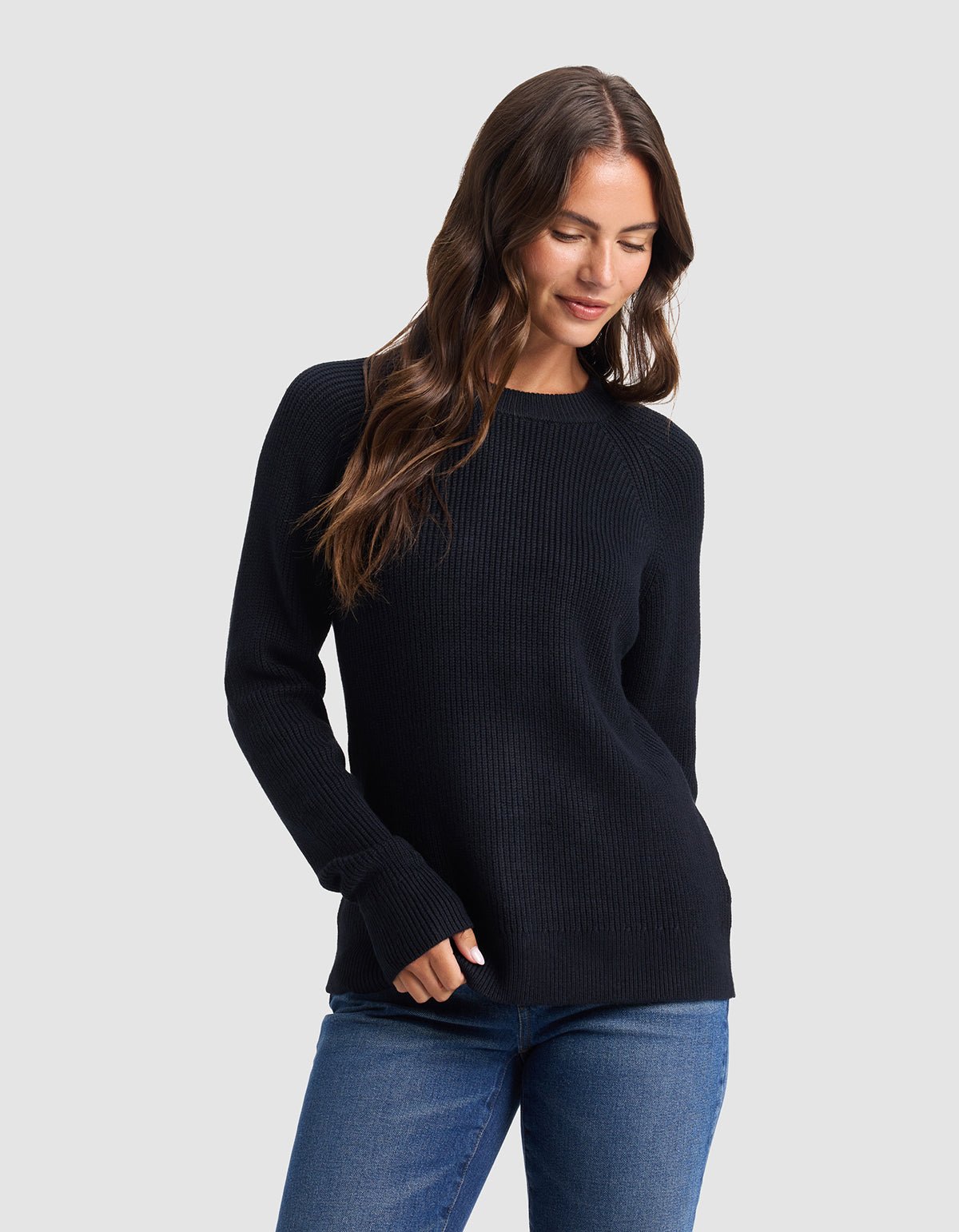 A woman with long brown hair models the Cozy Earth Women's Classic Crewneck, wearing blue jeans and standing against a light gray background, looking down with a slight smile and one hand in her pocket. 