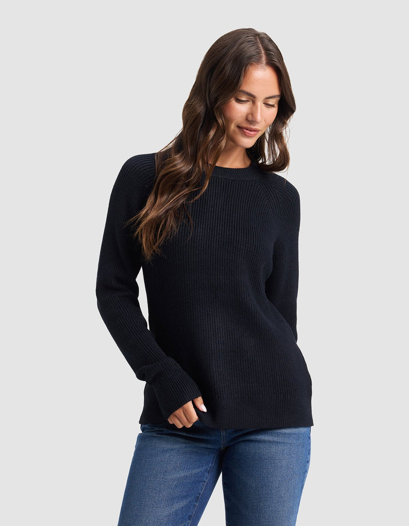 A woman with long brown hair models the Cozy Earth Women's Classic Crewneck, wearing blue jeans and standing against a light gray background, looking down with a slight smile and one hand in her pocket.