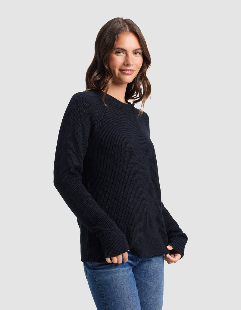 A woman with wavy brown hair smiles softly while wearing the Cozy Earth Women's Classic Crewneck in black and blue jeans, standing against a plain light gray background with her hands relaxed by her sides.